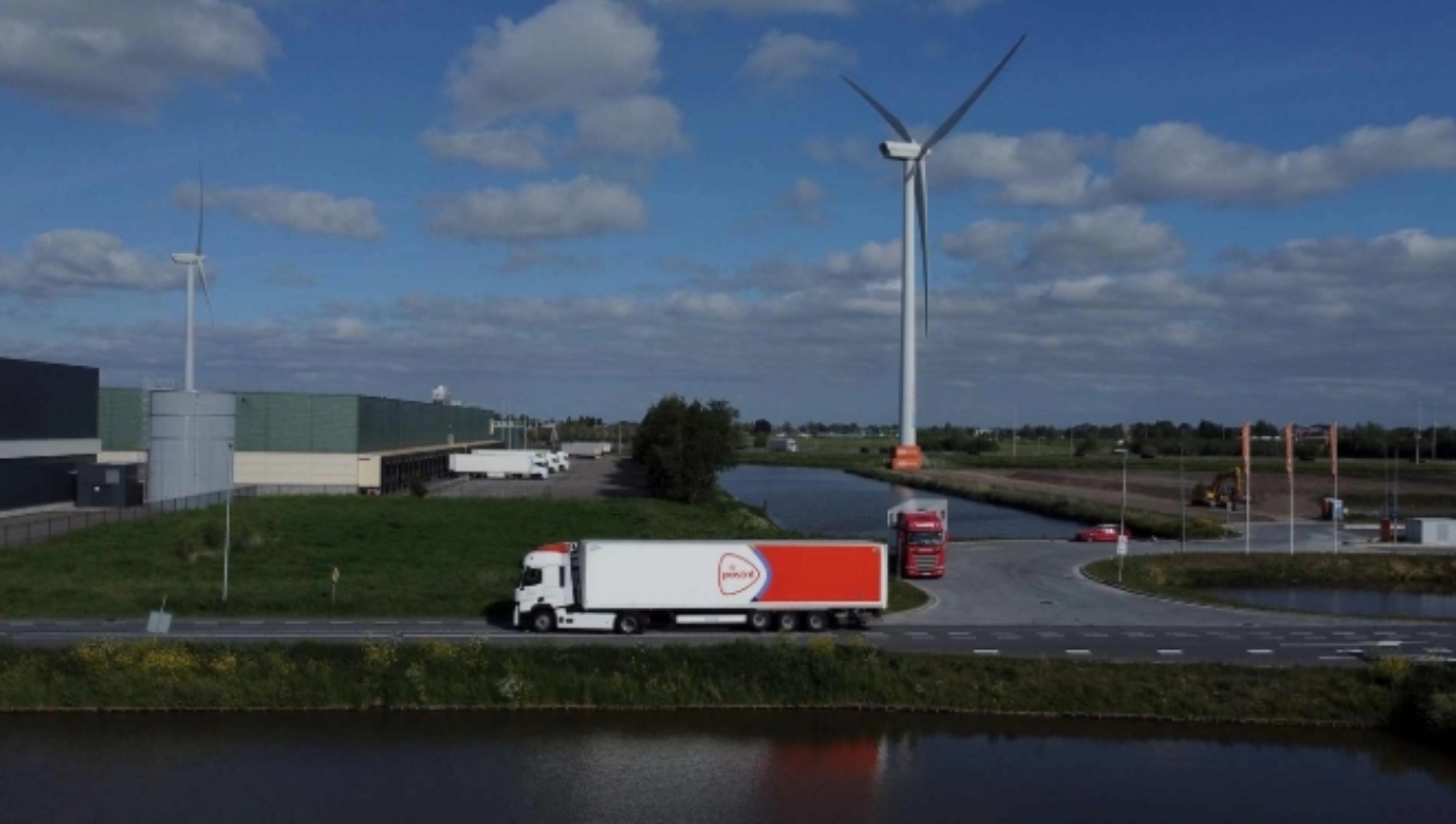 A white and orange PostNL truck drives past a warehouse and wind turbines near a canal.