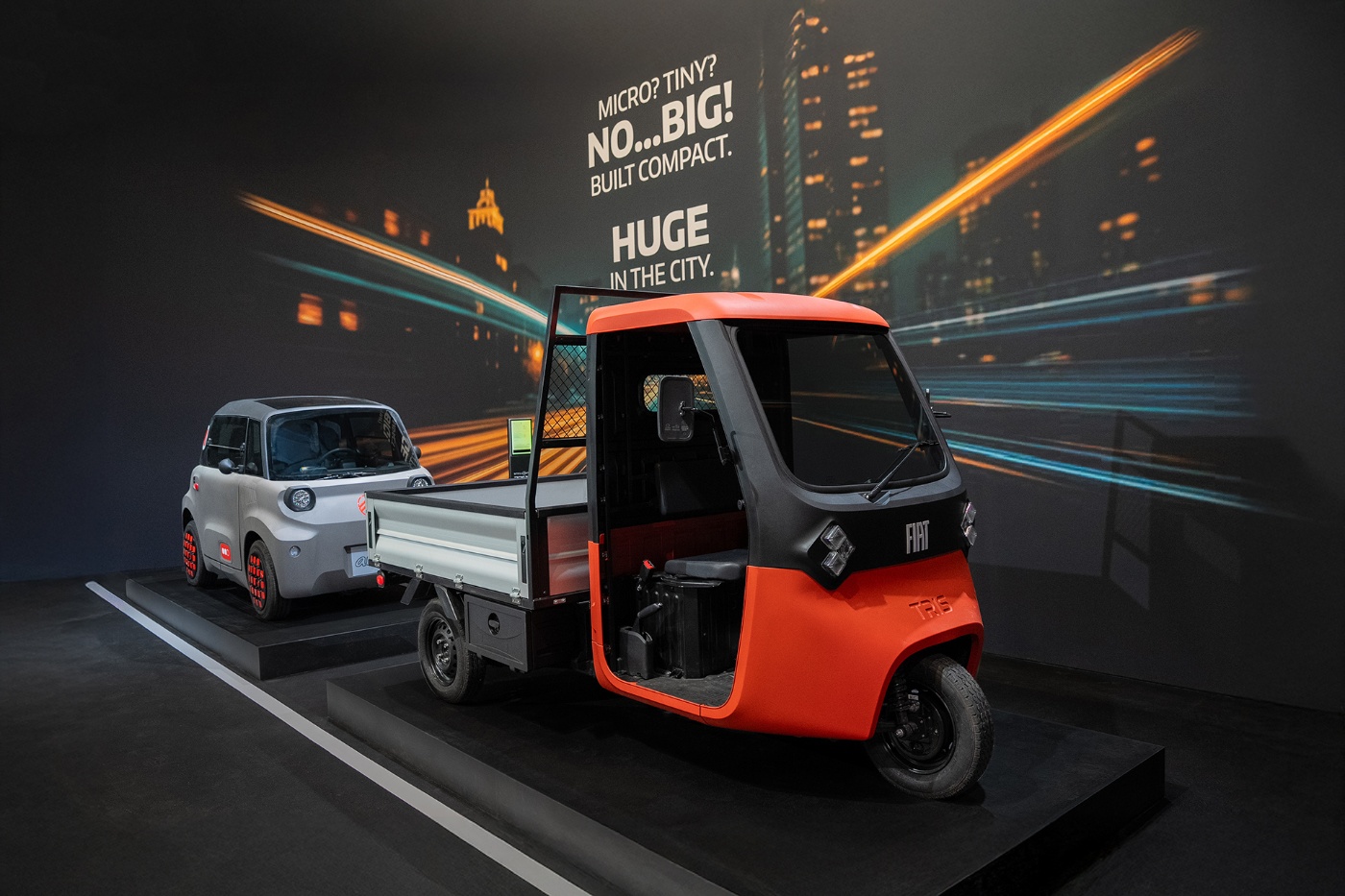 Ein roter Fiat-Minilaster und ein kompaktes Elektroauto stehen vor einem städtischen Hintergrund.
