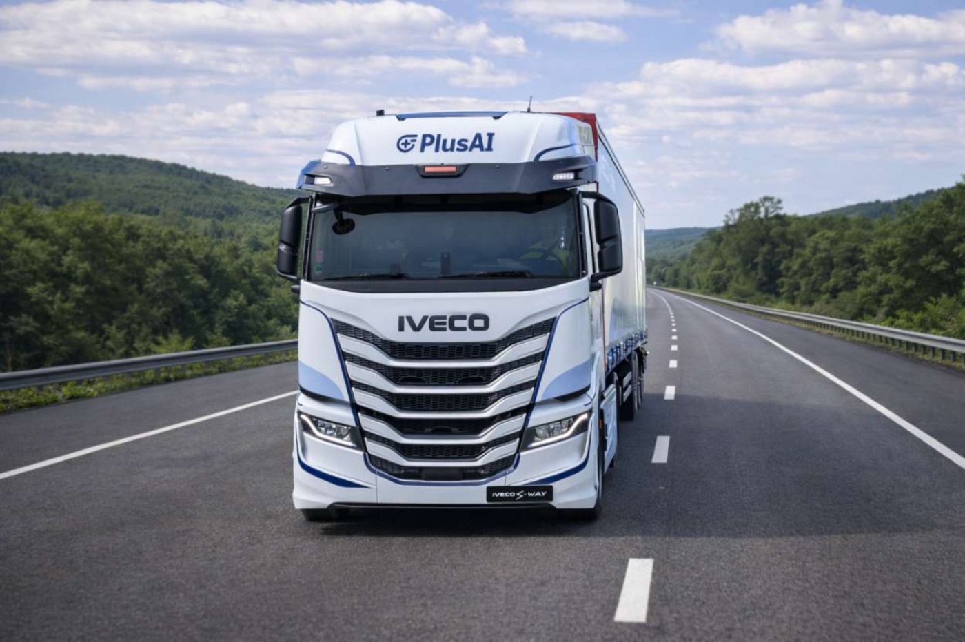 Ein weißer Iveco-PlusAI-Lkw fährt auf einer leeren Autobahn zwischen grünen Hügeln.