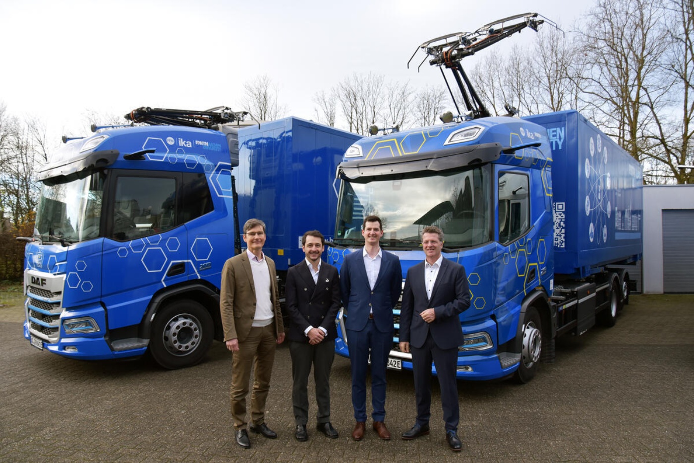 Vier Männer stehen vor zwei blauen E-Lkw mit Stromabnehmern und Technikgrafiken.