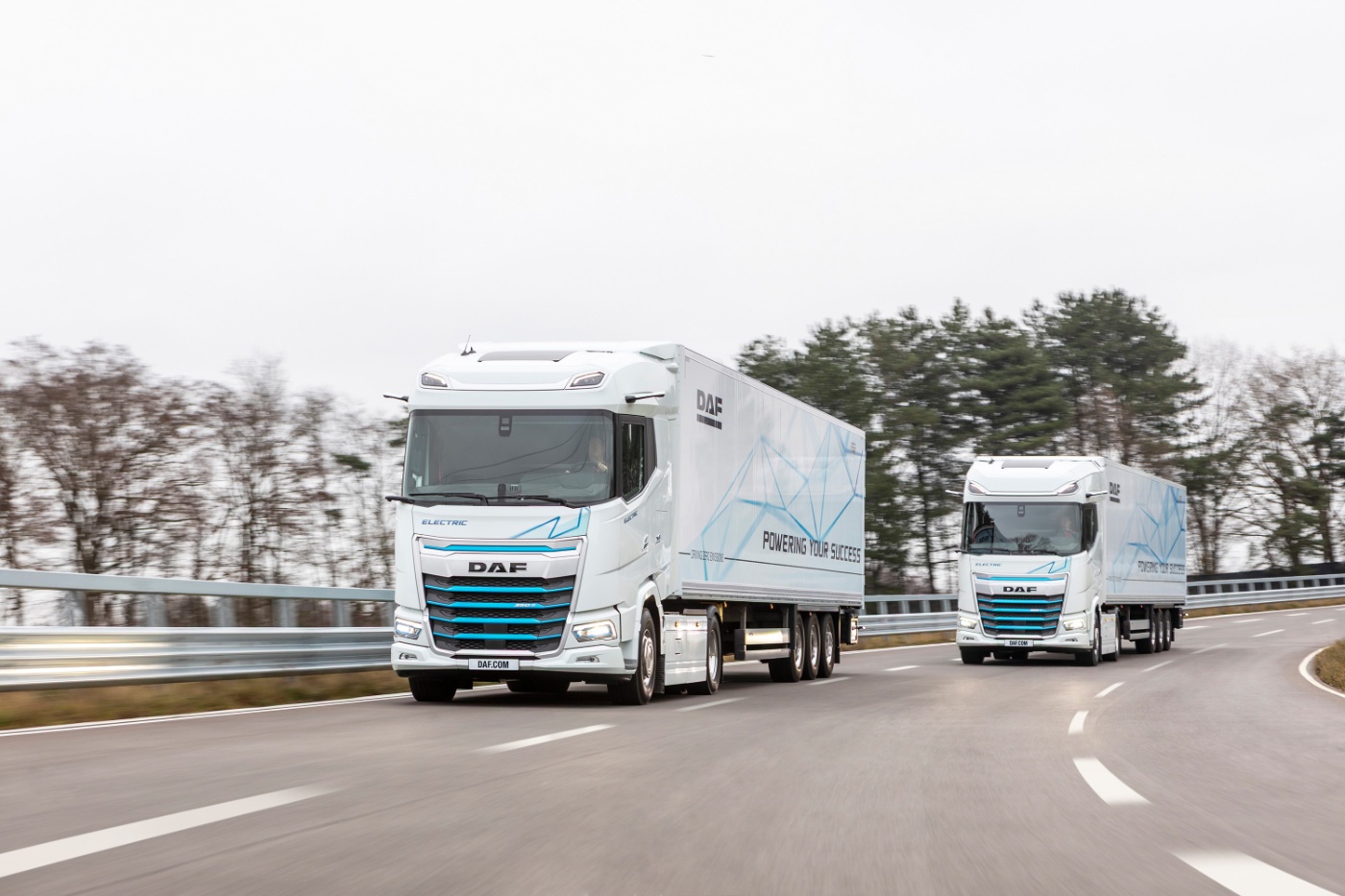 Two electric DAF trucks driving on a curved road with trees in the background.