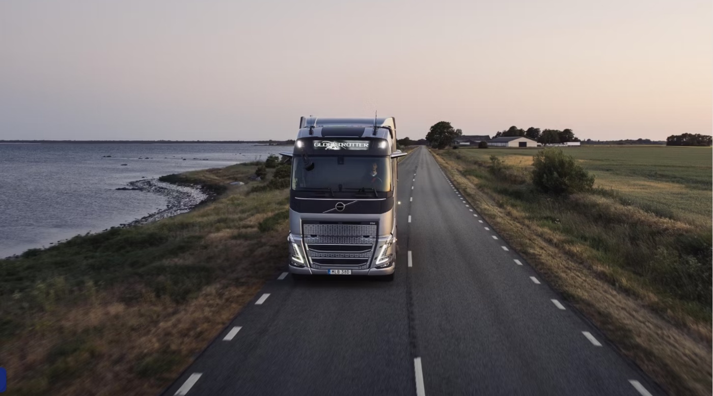 A Volvo truck drives along a coastal road at sunset, passing fields and buildings.