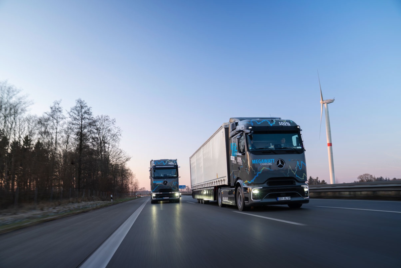 Two electric Mercedes-Benz trucks with "Megawatt Charging" branding drive on a highway at dusk.