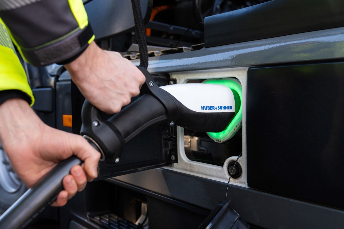 A person plugs a HUBER+SUHNER charging cable into an electric truck with green light indicator.