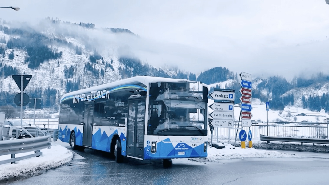 Electric bus driving on a snowy mountain road near directional signs.