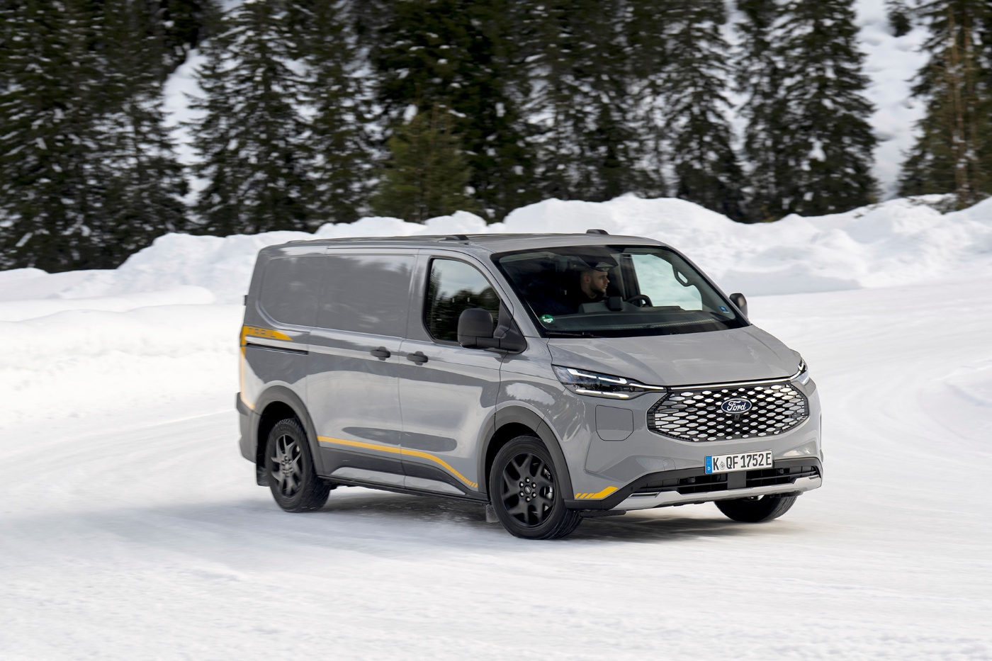 Grauer elektrischer Ford-Transporter fährt auf verschneiter Straße vor Wald.