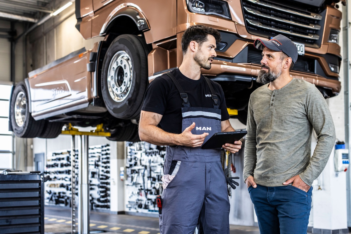 Two mechanics talk in a workshop while a truck is lifted on a car hoist.