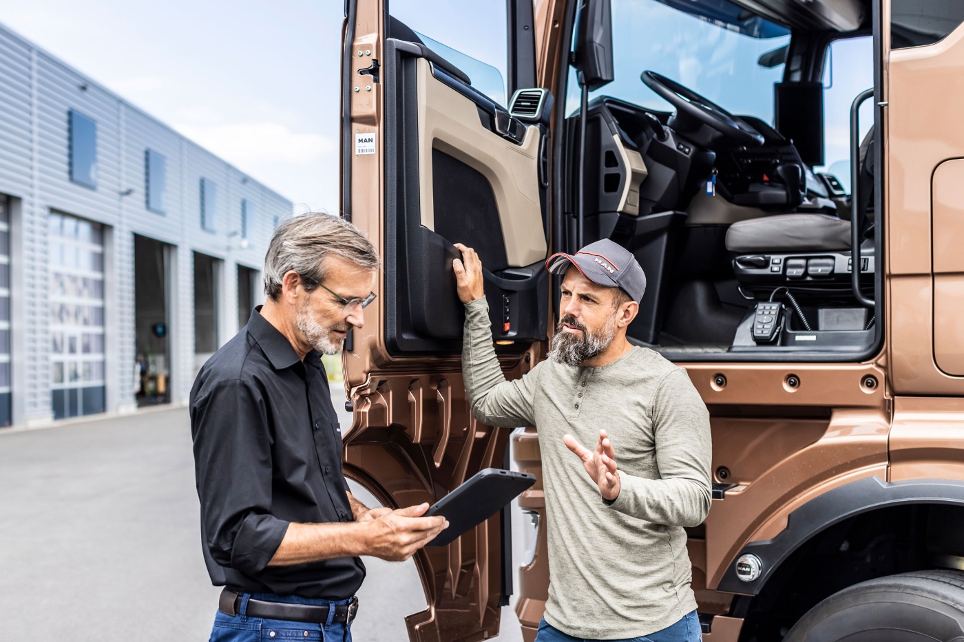 Two men stand by a truck cab, discussing while one holds a clipboard.