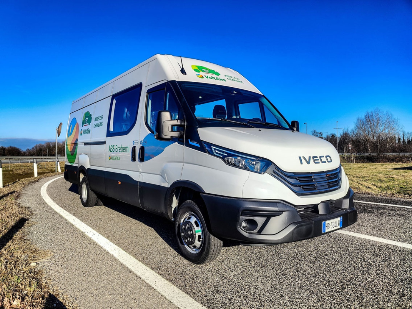 A white Iveco van drives on a curved road under a clear blue sky.