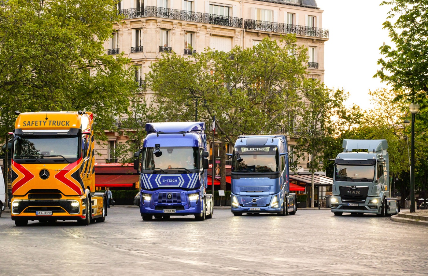 Four trucks, including electric models, parked side by side in a city square.