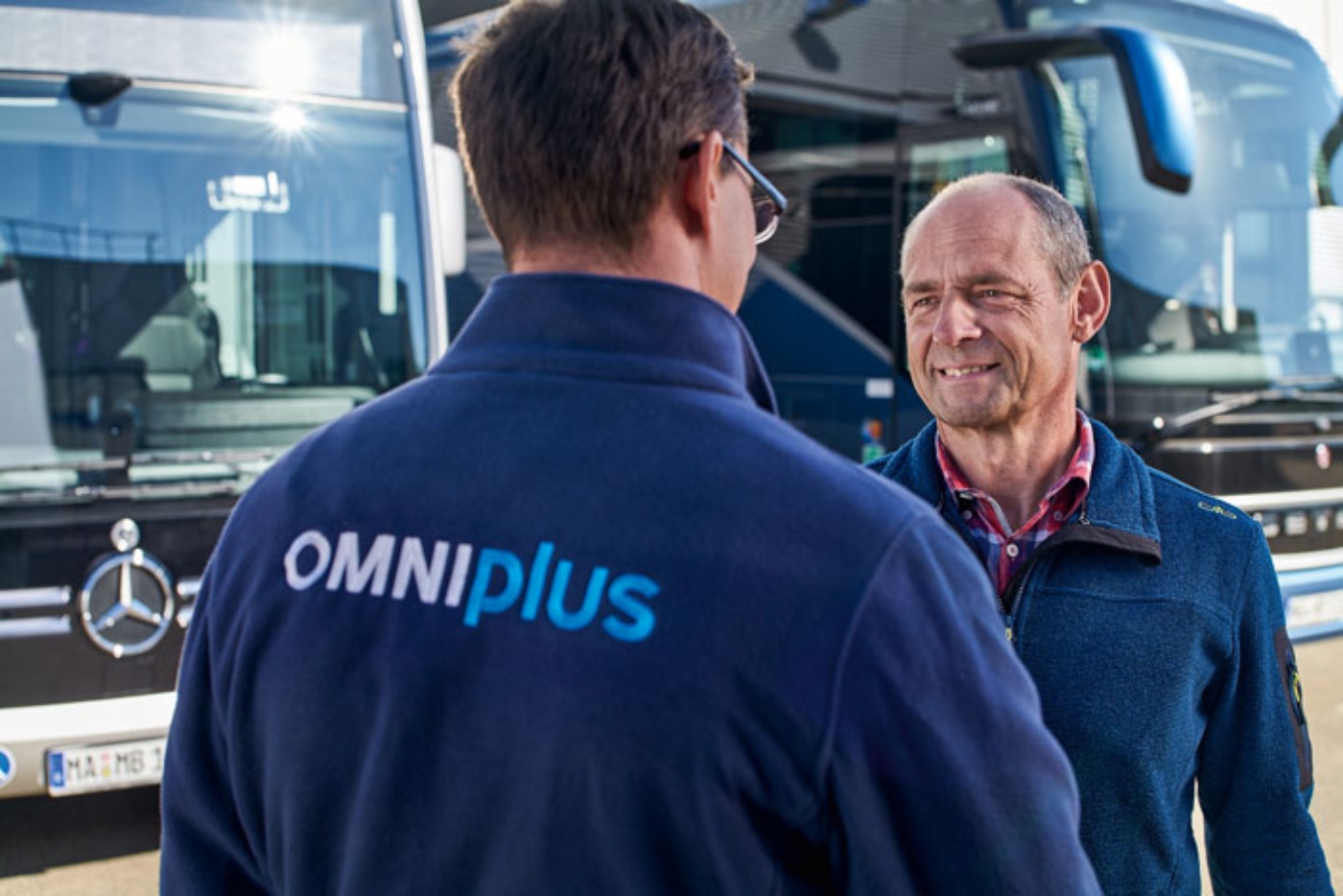 Two men talking in front of buses, one wearing a jacket with Omniplus logo.