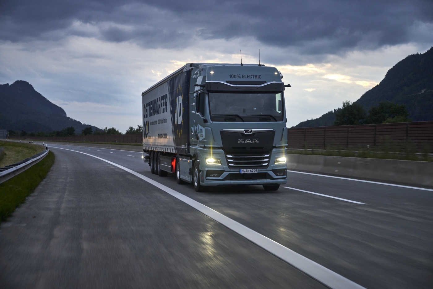Truck driving on wet highway between mountains under cloudy sky.