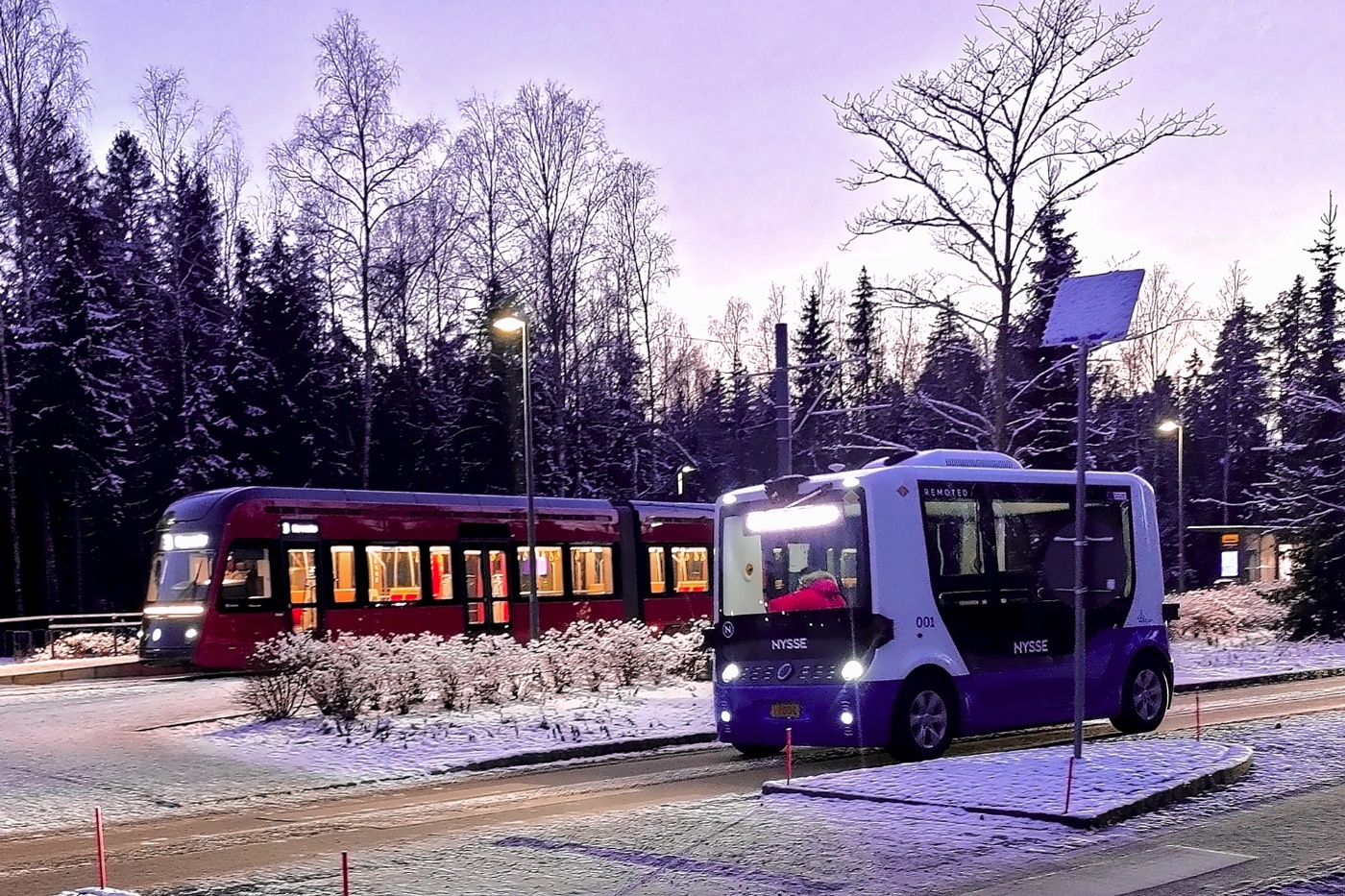 Autonomous shuttle bus driving beside a tram on a snowy street.