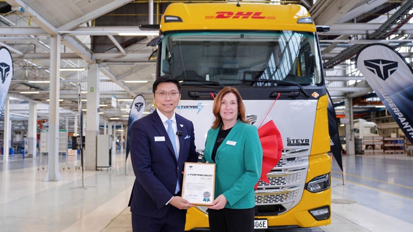 Two people holding a certificate in front of a DHL electric truck in a production hall.