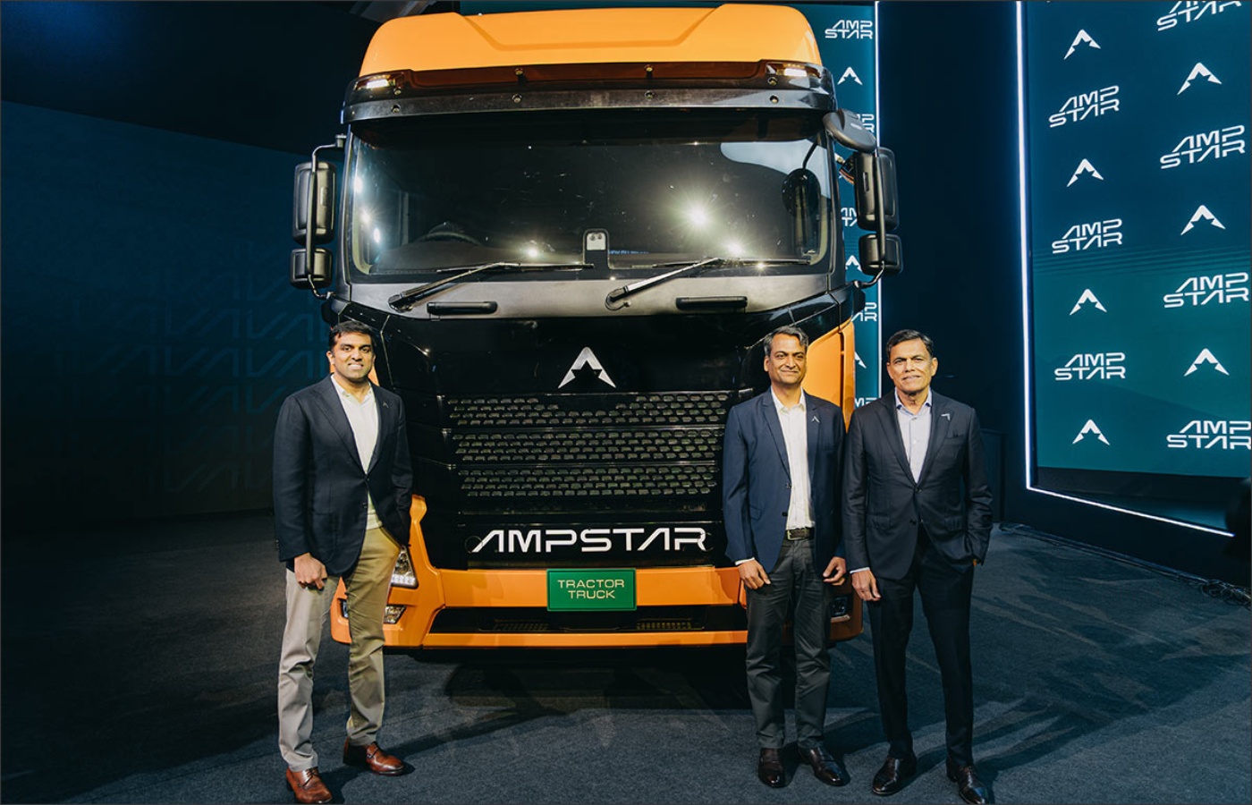 Three men stand in front of an orange and black AMPSTAR tractor truck.