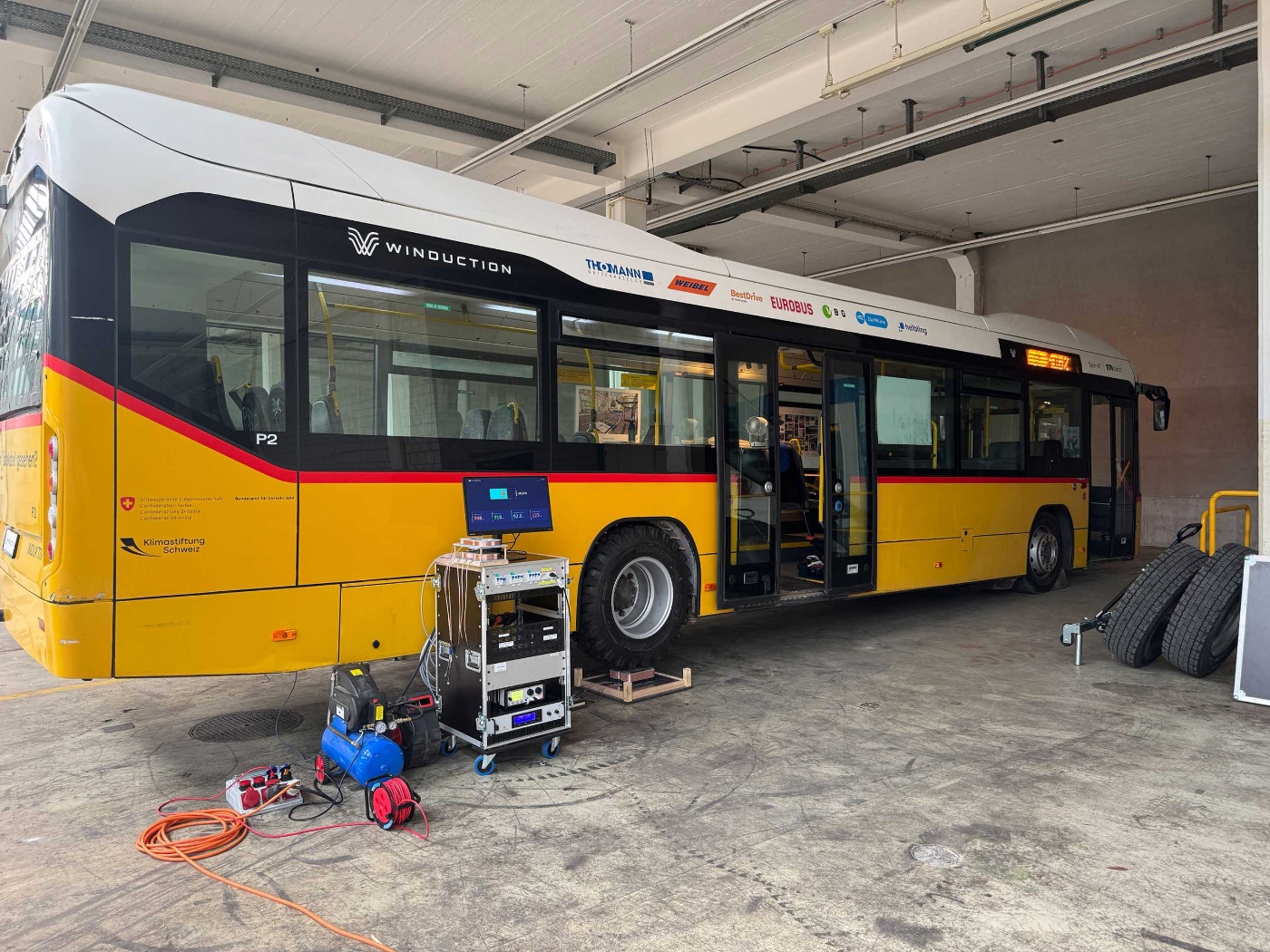 Yellow electric bus in a workshop with open doors and technical equipment in front of it.