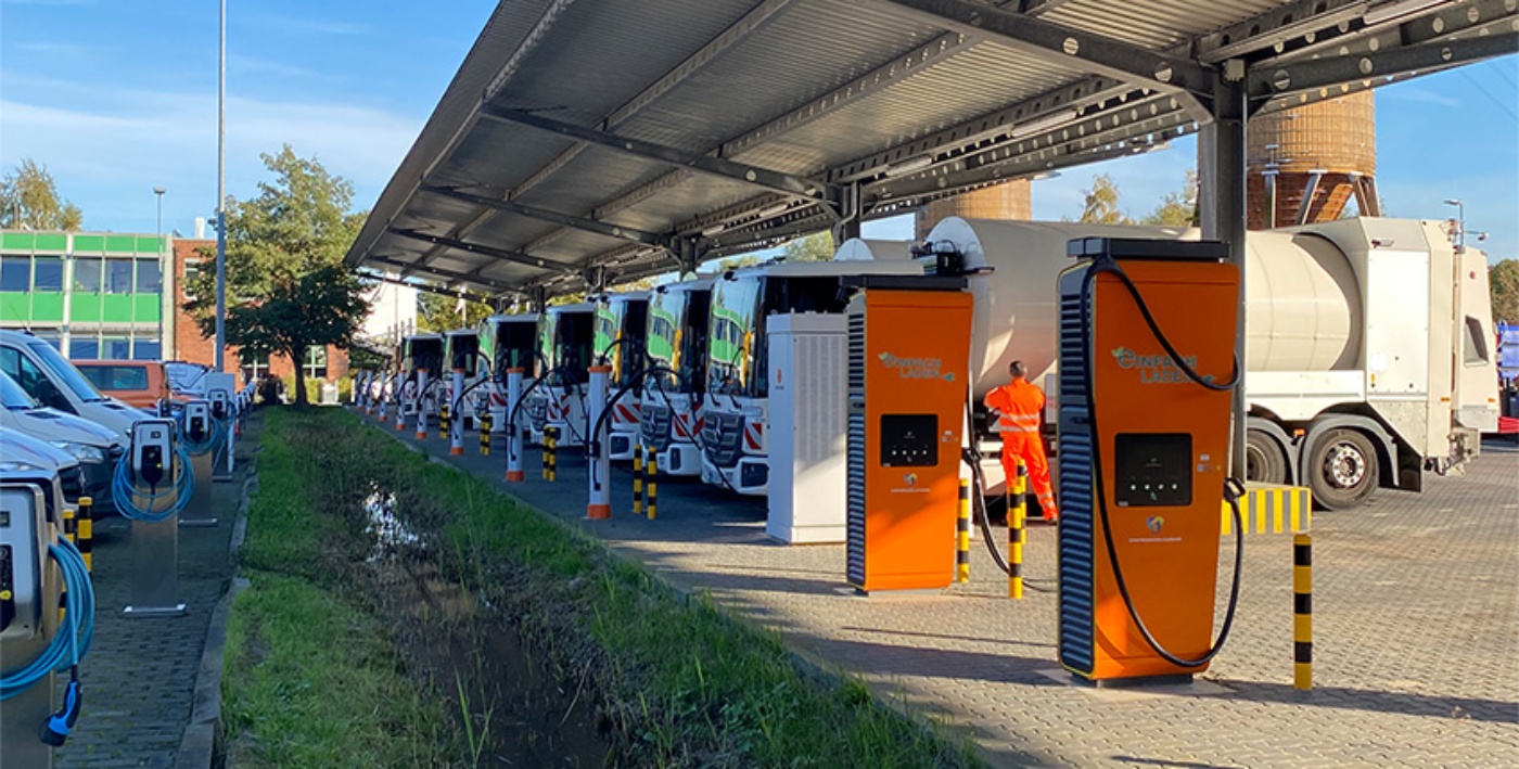 Orange charging stations beneath a solar canopy at a parking area with parked trucks.