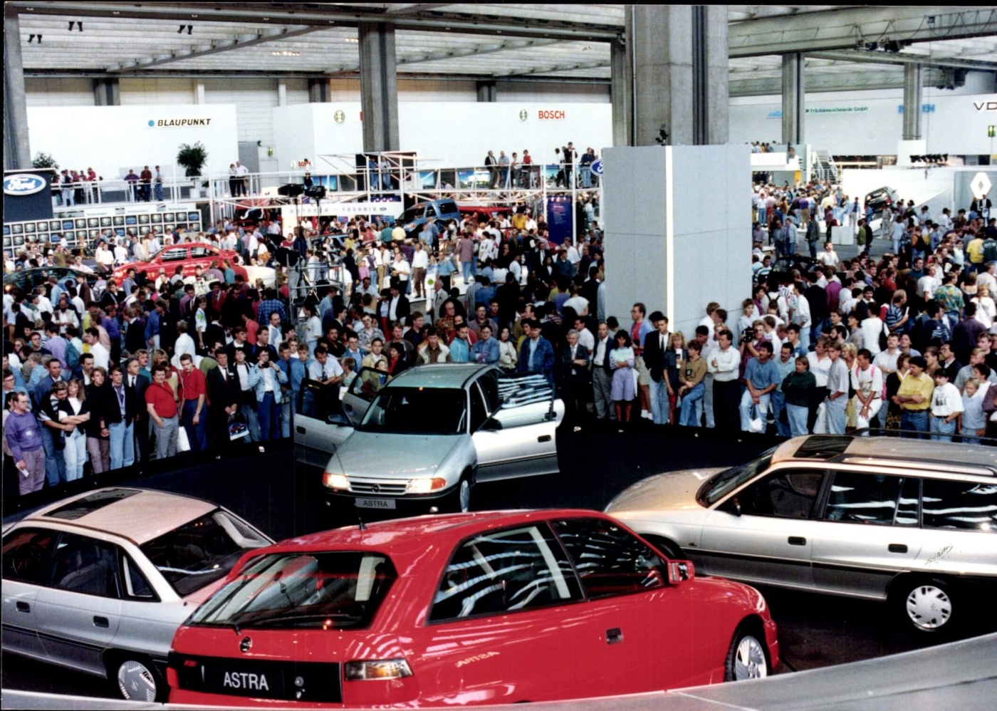 A large crowd gathers in an exhibition hall with several cars displayed at an IAA event.