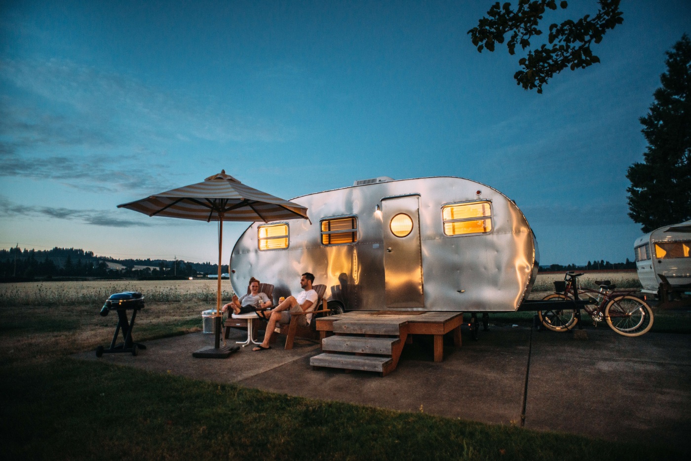 Camping trailer at dusk with lights on and people sitting outside under an umbrella.